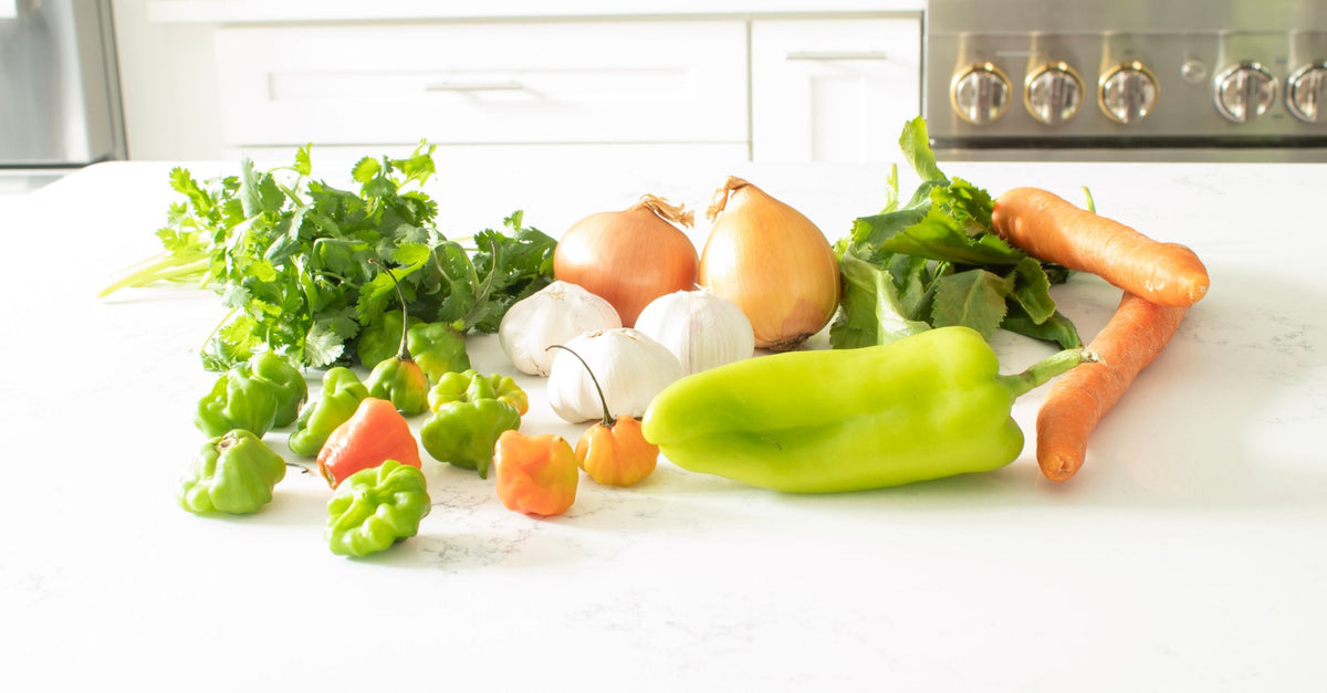 Sofrito ingredients on a kitchen counter: cilantro, ají dulce peppers, garlic, onions, recao, a long green pepper and carrots