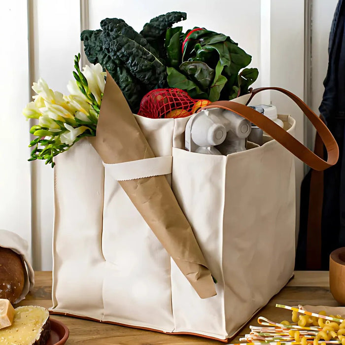 Cream canvas market tote standing on a wooden counter, packed with kale, chard, tomatoes in a red mesh bag and a paper-wrapped bouquet, tan handles over the side