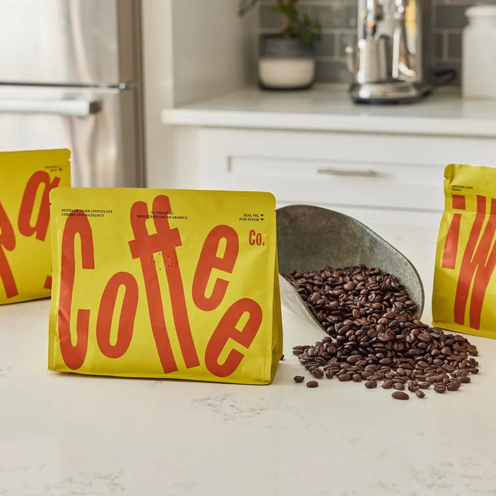 Three yellow Wepa Coffee bags on a kitchen counter with roasted beans spilling from a metal scoop